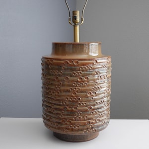 May include: A brown ceramic table lamp with a textured, bubble-like pattern. The lamp has a brass neck and a light fixture at the top. The base is a darker shade of brown.