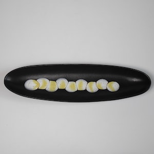 May include: A long, oval black ceramic dish with a series of white and yellow decorative elements. The dish is centered against a plain white background. The decorative elements are arranged in a line. The dish is likely for serving food.