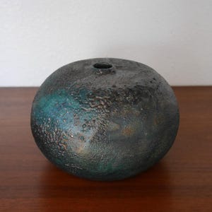 May include: A dark green and black ceramic vase with a textured surface. The vase is round and has a small opening at the top.