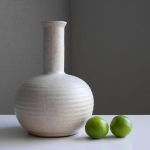 May include: A cream-colored ceramic vase with a round body and a long, slender neck. The vase has horizontal ridges around the body. Two green limes sit to the right of the vase. The vase and limes are on a white surface.