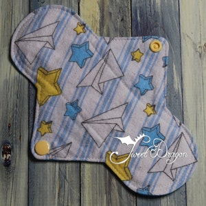 May include: A reusable cloth menstrual pad with a white background and a pattern of blue stars, yellow stars, and paper airplanes. The pad has two yellow snaps on the sides.