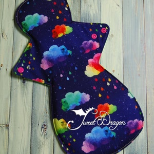 May include: A reusable cloth menstrual pad with a dark blue background and a colorful rainbow cloud pattern. The pad has a green trim and pink snaps. The text "Sweet Dragon" is printed in white on the pad.
