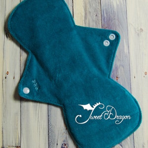 May include: A teal cloth menstrual pad with a curved shape and snap closures. The pad has the words "Sweet Dragon" in a cursive font with a dragon graphic. The pad is made of a soft, absorbent material.