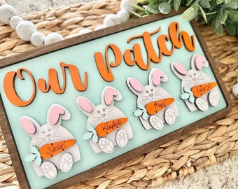 Peut inclure: Une pancarte en bois avec le texte "Our Patch" en lettres orange. Il y a quatre figurines de lapins gris tenant des carottes avec des noms écrits dessus : Justin, Kaylee, Myla et Blanjer.