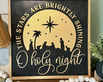 May include: Wooden Christmas sign with a black background and gold accents. The sign features the text "The Stars Are Brightly Shining, O Holy Night" and a silhouette of the nativity scene.