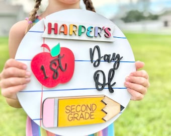 May include: A round, white wooden sign with the name "Harper's" in colorful letters. The sign features a red apple with "1st", the words "Day Of", and a yellow pencil with "Second Grade". The sign is held by a young girl.