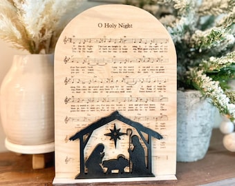 Könnte beinhalten: Eine hölzerne Weihnachtskrippe mit einer schwarzen Silhouette von Maria, Josef und dem Jesuskind. Die Szene ist vor dem Hintergrund eines Notenblattes mit dem Text des Weihnachtsliedes "O Holy Night" platziert.