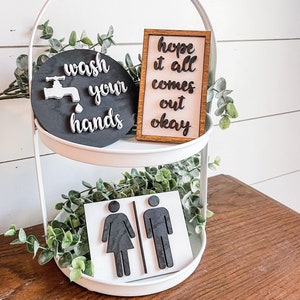 May include: A white tiered tray with three signs. The top sign is black with a white faucet and the text "wash your hands". The middle sign is wood with the text "hope it all comes out okay". The bottom sign is white with black silhouettes of a woman and a man.