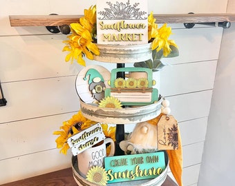 Puede incluir: Una bandeja de tres niveles decorada con girasoles, gnomos y letreros. El nivel superior tiene un letrero que dice "Sunflower Market". El nivel intermedio tiene un camión verde con girasoles y un gnomo. El nivel inferior tiene una taza, un letrero que dice "Good Morning", un letrero que dice "Sunflower Maze", y un letrero que dice "Create Your Own Sunshine".