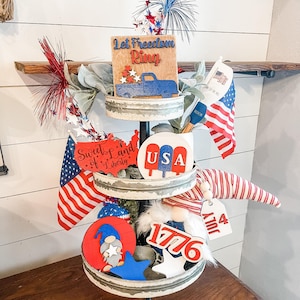 Puede incluir: Una bandeja de tres niveles decorada con adornos patri&oacute;ticos rojo, blanco y azul. El nivel superior presenta un letrero de madera que dice "Let Freedom Ring" con una camioneta roja, blanca y azul. El nivel intermedio presenta un letrero de madera que dice "Sweet Land of Liberty" y un letrero de madera que dice "USA". El nivel inferior presenta un letrero de madera que dice "1776" y un letrero de madera que dice "4th July".