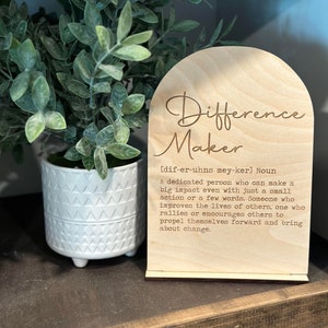 May include: Wooden sign with the words "Difference Maker" and a definition of the word. The sign is on a wooden stand and has a light brown color.