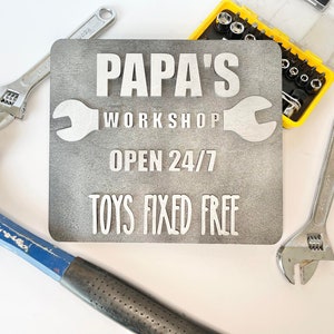May include: A gray wooden sign with white text that reads "Papa's Workshop Open 24/7 Toys Fixed Free". The sign is surrounded by tools, including wrenches and a hammer.