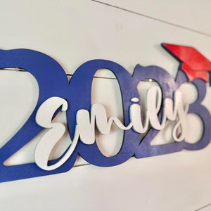 Grad Sign shown with 2023 with the 2023 in Royal Blue, The Hat in Red and the name in white by walnut + birch