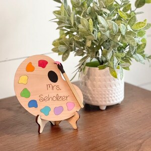 May include: Wooden paint palette with a paintbrush, standing on a small easel. The palette has a rainbow of colours and the text "Mrs. Scholz" is written on it.