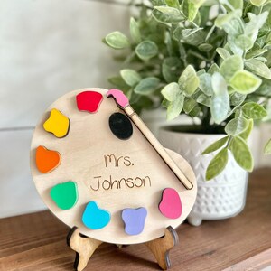 May include: A wooden paint palette with a paintbrush and colorful paint dots. The palette has the text "mrs. Johnson" written on it.