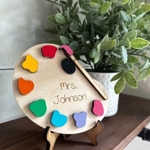 May include: A wooden paint palette with a paintbrush and colorful paint dots. The palette has the text "Mrs. Johnson" written on it.