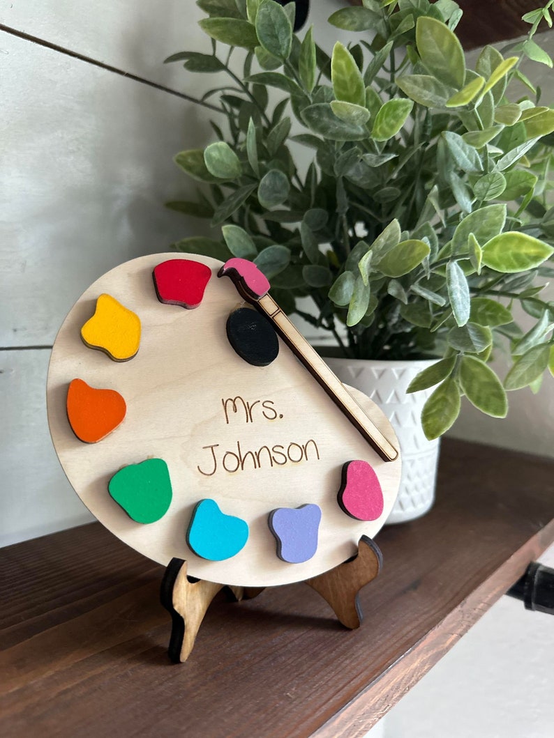 May include: A wooden paint palette with a paintbrush and colorful paint dots. The palette has the text "Mrs. Johnson" written on it.