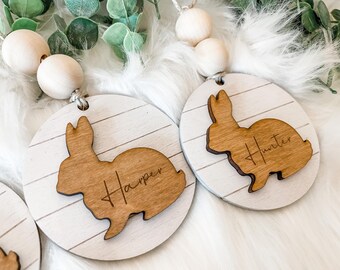 May include: Two wooden Easter bunny ornaments with personalized names, "Harper" and "Hunter", on a white background. The ornaments are attached to a string of wooden beads.
