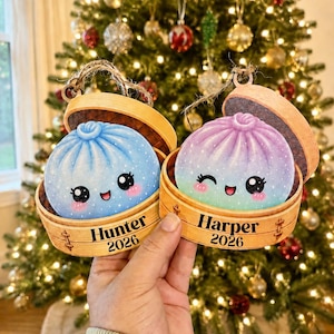 May include: Two round wooden ornaments shaped like bamboo steamer baskets containing colorful dumplings. One dumpling is blue, the other is a purple-to-green gradient. Both feature smiling faces and are personalized with the names Hunter and Harper and the year 2026.
