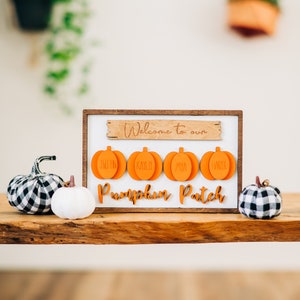 May include: Welcome to our Pumpkin Patch sign with four orange pumpkin-shaped ornaments with names on them. The sign is on a wooden table with two small decorative pumpkins.