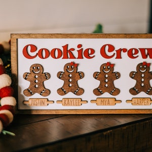 May include: A wooden sign with the words "Cookie Crew" in red letters. There are four gingerbread people cutouts with names below each: Justin, Kaylee, Mya, and Harper.
