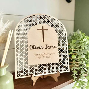 May include: A wooden sign with an arched top and a decorative lattice design. The sign features a cross and the text "Oliver James, First Holy Communion, April 26, 2026." It is supported by a wooden stand and placed on a wooden shelf.