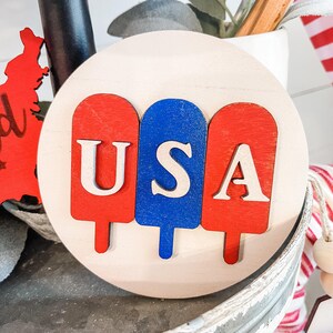 Puede incluir: Un letrero redondo de madera con tres piezas de madera en forma de paleta de helado pintadas de rojo, blanco y azul. Las letras "USA" est&aacute;n cortadas en la madera y pintadas de blanco.