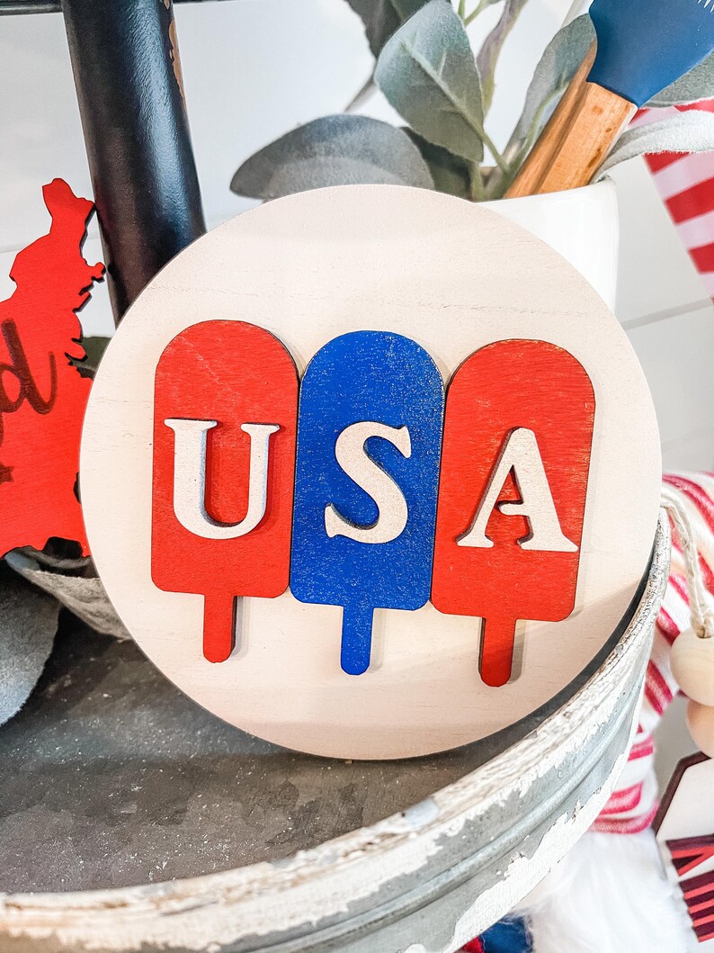 Puede incluir: Un letrero redondo de madera con tres piezas de madera en forma de paleta de helado pintadas de rojo, blanco y azul. Las letras "USA" est&aacute;n cortadas en la madera y pintadas de blanco.