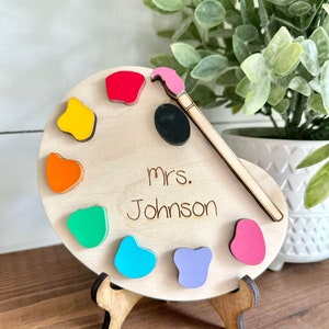 May include: Wooden paint palette with a paintbrush and colorful paint drops. The palette has the name "mrs. Johnson" written on it.