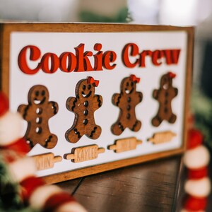 May include: A wooden sign with the words "Cookie Crew" in red letters. There are four gingerbread people cutouts with names below each: Austin, Kaylee, Micah, and Noah.
