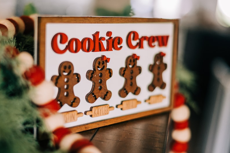 May include: A wooden sign with the words "Cookie Crew" in red letters. There are four gingerbread people cutouts with names below each: Austin, Kaylee, Micah, and Noah.