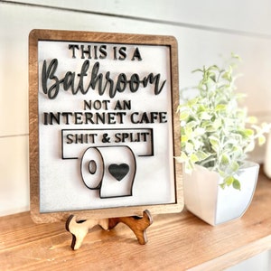 May include: A wooden sign with the text "This is a Bathroom Not an Internet Cafe Shit & Split" and a cartoon toilet paper roll with a heart on it. The sign is on a small wooden easel.