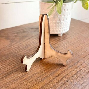 May include: A wooden picture stand with a light brown finish. The stand has a triangular shape with three legs.