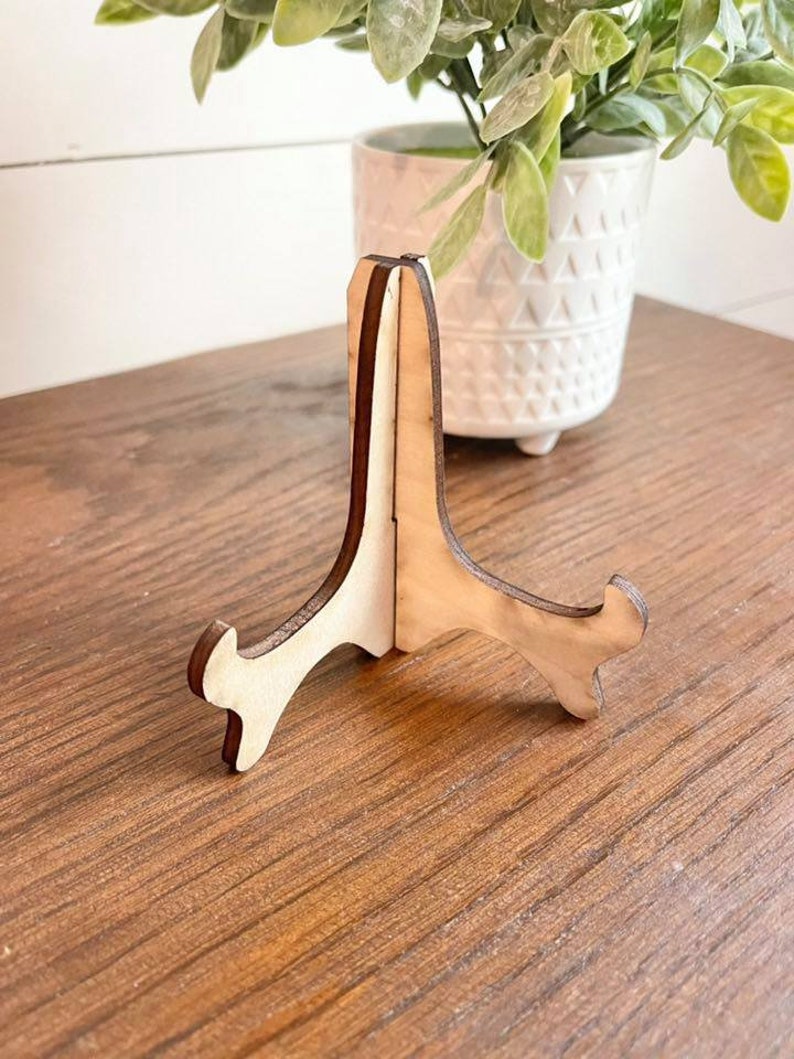 May include: A wooden picture stand with a light brown finish. The stand has a triangular shape with three legs.