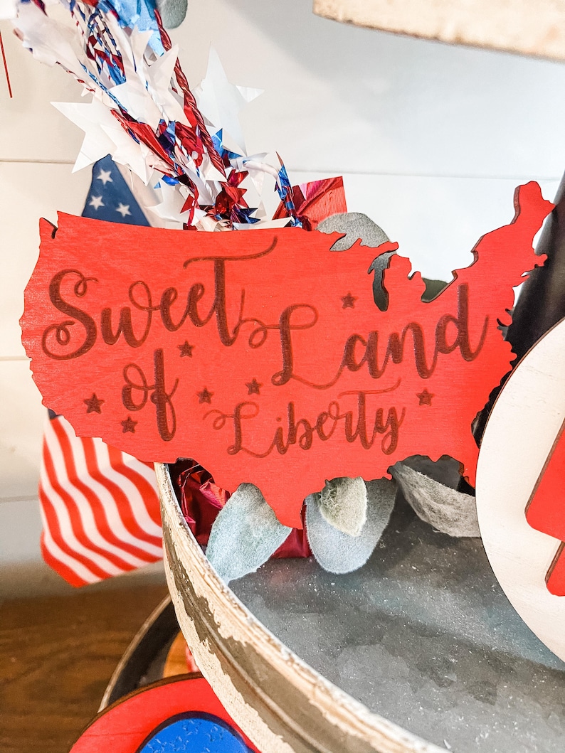 Puede incluir: Corte de madera rojo en forma de los Estados Unidos con el texto "Sweet Land of Liberty" en letras marrones.