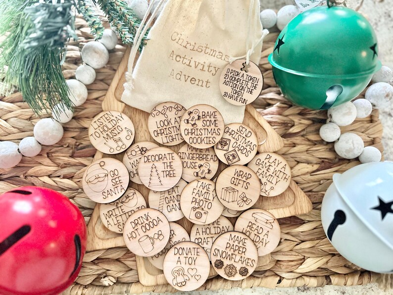 May include: A set of 20 wooden Christmas activity tokens with various holiday-themed illustrations and text, such as "Listen to Holiday Music", "Bake Some Christmas Cookies", and "Donate a Toy". The tokens are scattered on a woven basket with a white and green jingle bell and a red jingle bell.