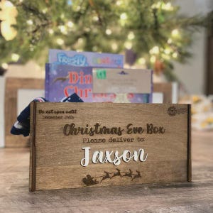 Op de afbeelding: Een rechthoekige houten kerstavonddoos met gegraveerde tekst Do not open until December 24th, Christmas Eve Box en Please deliver to: Jaxson. De doos is voorzien van een lasergesneden silhouet van de kerstman in een arrenslee met rendieren.