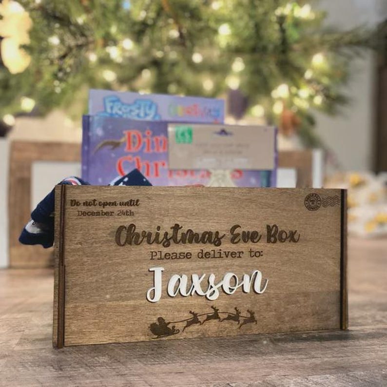 Op de afbeelding: Een rechthoekige houten kerstavonddoos met gegraveerde tekst Do not open until December 24th, Christmas Eve Box en Please deliver to: Jaxson. De doos is voorzien van een lasergesneden silhouet van de kerstman in een arrenslee met rendieren.