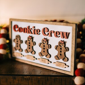May include: A wooden sign with the words "Cookie Crew" in red letters. There are three gingerbread people cutouts with the names "Kaylee", "Mya", and "Harper" below them.