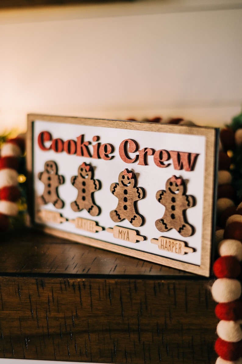 May include: A wooden sign with the words "Cookie Crew" in red letters. There are three gingerbread people cutouts with the names "Kaylee", "Mya", and "Harper" below them.