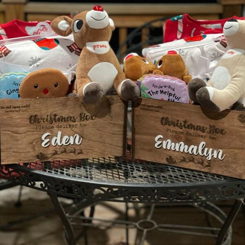 Op de afbeelding: Twee houten kerstkisten met de tekst "Christmas Box Please deliver to:" en de namen "Eden" en "Emmalyn" in wit schrift. Elke kist bevat pluche rendieren, witte stoffen items met sneeuwvlokpatronen en kleine kleurrijke bordjes.