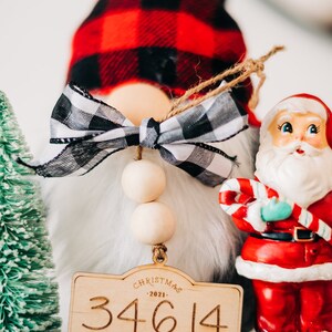 May include: A Christmas gnome with a red and black plaid hat, a white beard, and a wooden sign that says "Christmas 2021 34614 Home".