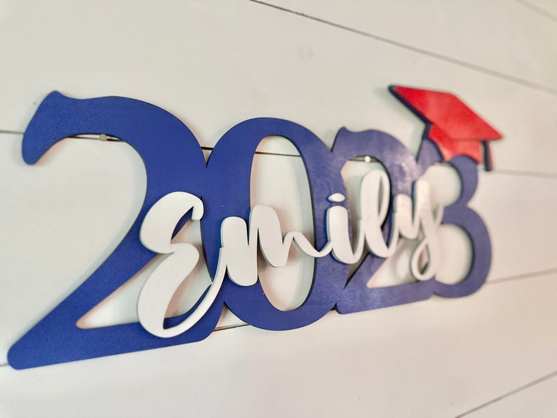 Grad Sign shown with 2023 with the 2023 in Royal Blue, The Hat in Red and the name in white by walnut + birch