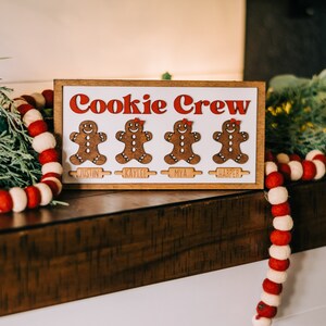 May include: A wooden sign with the text "Cookie Crew" in red letters. Four gingerbread people are displayed on the sign, each with a name below: Justin, Kaylee, Mya, and Harper.