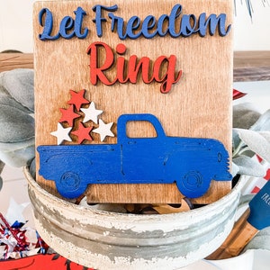 Puede incluir: Un letrero de madera con el texto "Let Freedom Ring" en letras azules y rojas. Debajo del texto hay un cami&oacute;n de madera azul con estrellas rojas y blancas en la caja.