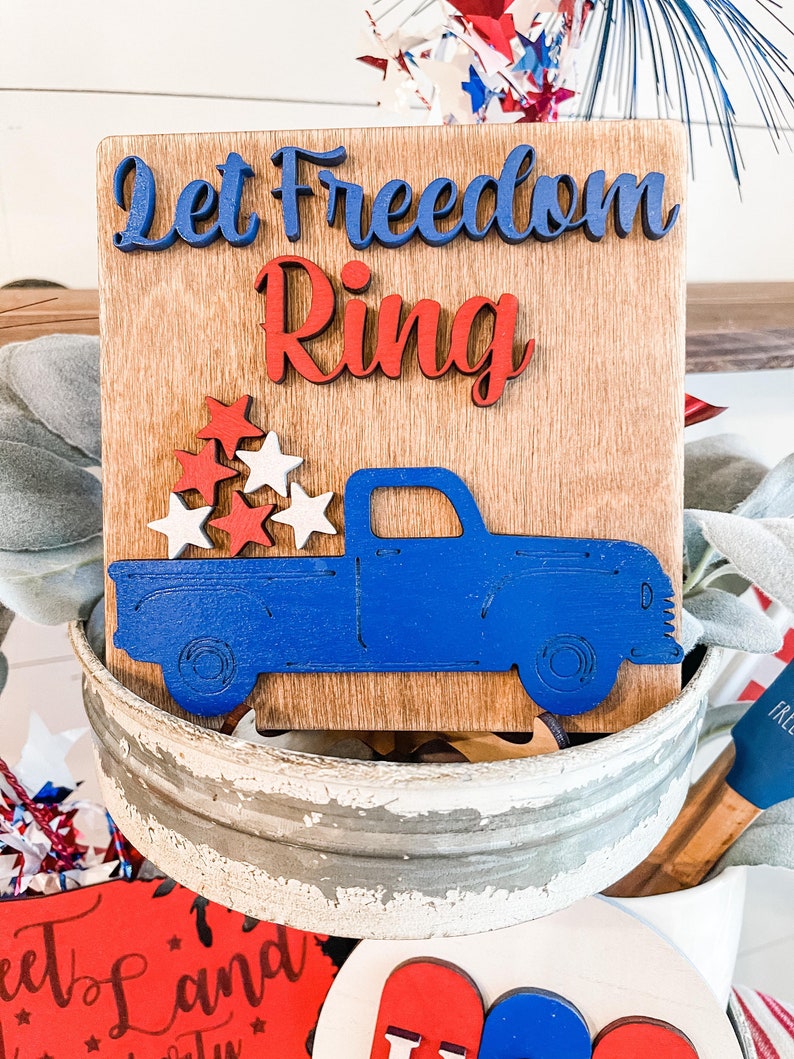 Puede incluir: Un letrero de madera con el texto "Let Freedom Ring" en letras azules y rojas. Debajo del texto hay un cami&oacute;n de madera azul con estrellas rojas y blancas en la caja.