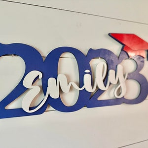 Grad Sign shown with 2023 with the 2023 in Royal Blue, The Hat in Red and the name in white by walnut + birch