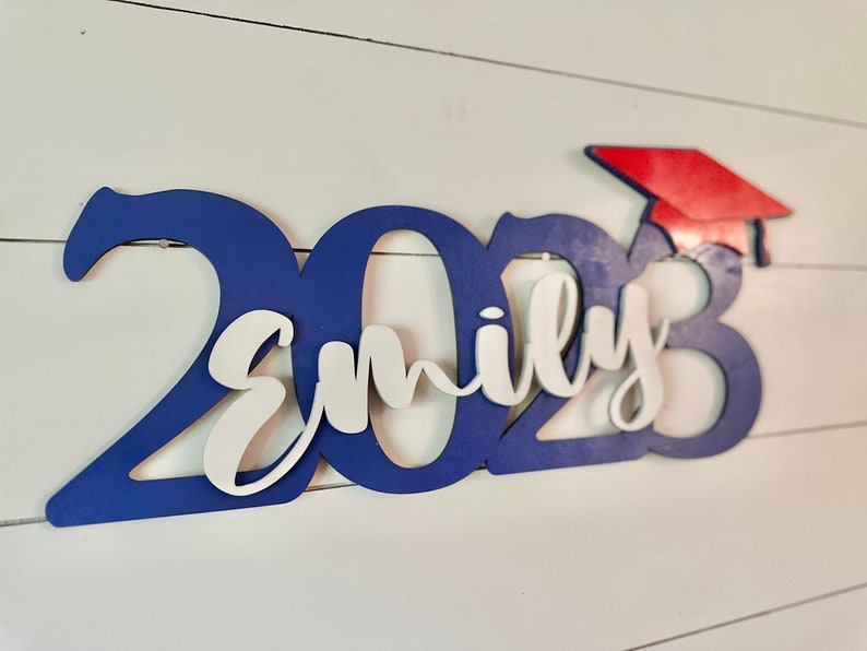 Grad Sign shown with 2023 with the 2023 in Royal Blue, The Hat in Red and the name in white by walnut + birch