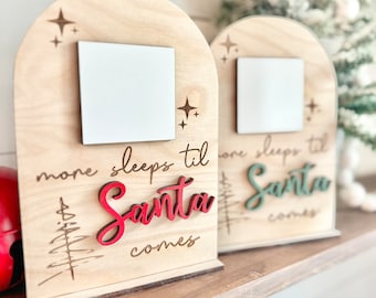 Könnte beinhalten: Zwei Holzschildchen mit einem weißen Quadrat in der Mitte. Die Schilder sind mit Sternen und einem Weihnachtsbaum verziert. Der Text auf den Schildern lautet "more sleeps til Santa comes" und "one sleeps til Santa comes". Das Wort "Santa" ist auf dem linken Schild rot und auf dem rechten Schild grün.