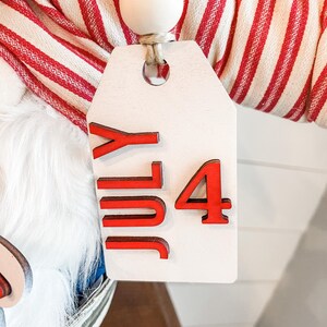 Puede incluir: Una etiqueta de madera blanca con letras rojas que dicen "July 4" colgando de una tela a rayas rojas y blancas. La etiqueta est&aacute; unida a una cuerda de cuentas de madera.
